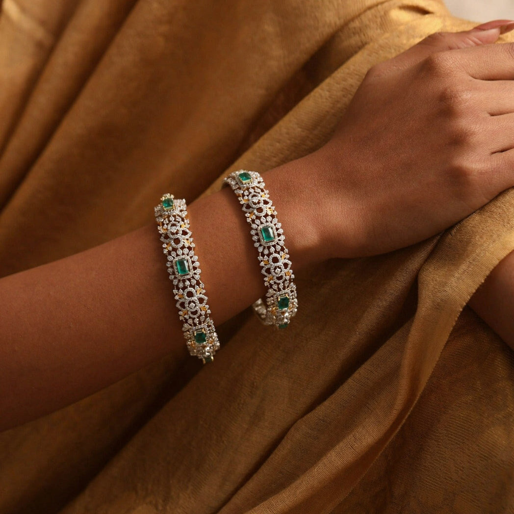 A pair of silver CZ bangles with green stones worn on the wrist against a saffron saree